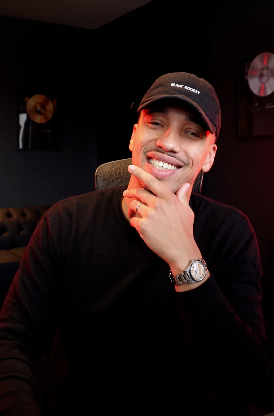 CJ sorrindo no estúdio, com discos de ouro e platina na parede